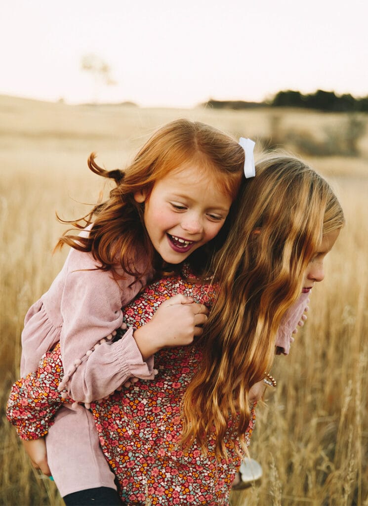 Family Photographer, an older her sister lets her younger sister ride on her back as she laughs in a dry meadow